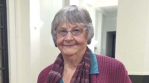 Head and shoulders photo of a lady wearing glasses wearing a maroon cardigan, standing in front of white walls.
