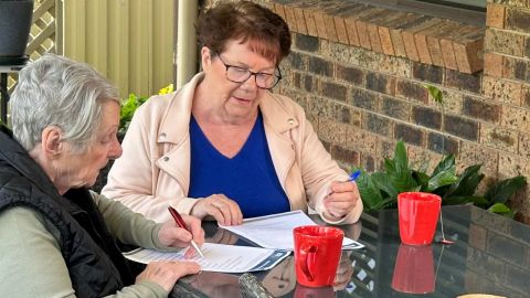 Two women sitting at a table, filling in documents with two red coffee cups on the table.