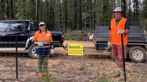 A woman and man wearing high vis and gloves stand behind a rural fence.
