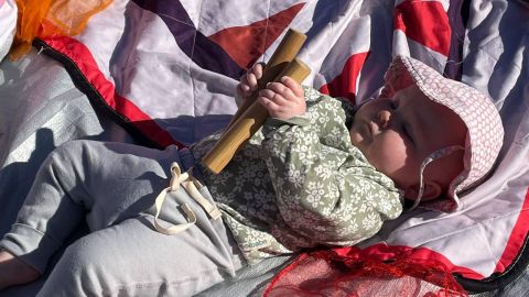 A baby lying on a blanket in the sun playing with a wooden musical instrument.