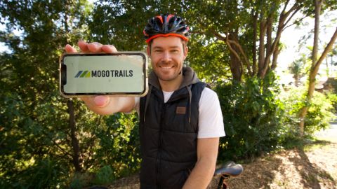 A man on a bike, wearing a bike helmet, holds up a smartphone on which is a logo with the words Mogo Trails.