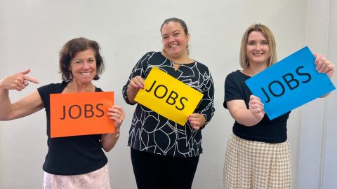 Three women smile at the camera each holding a sign that say 'jobs'