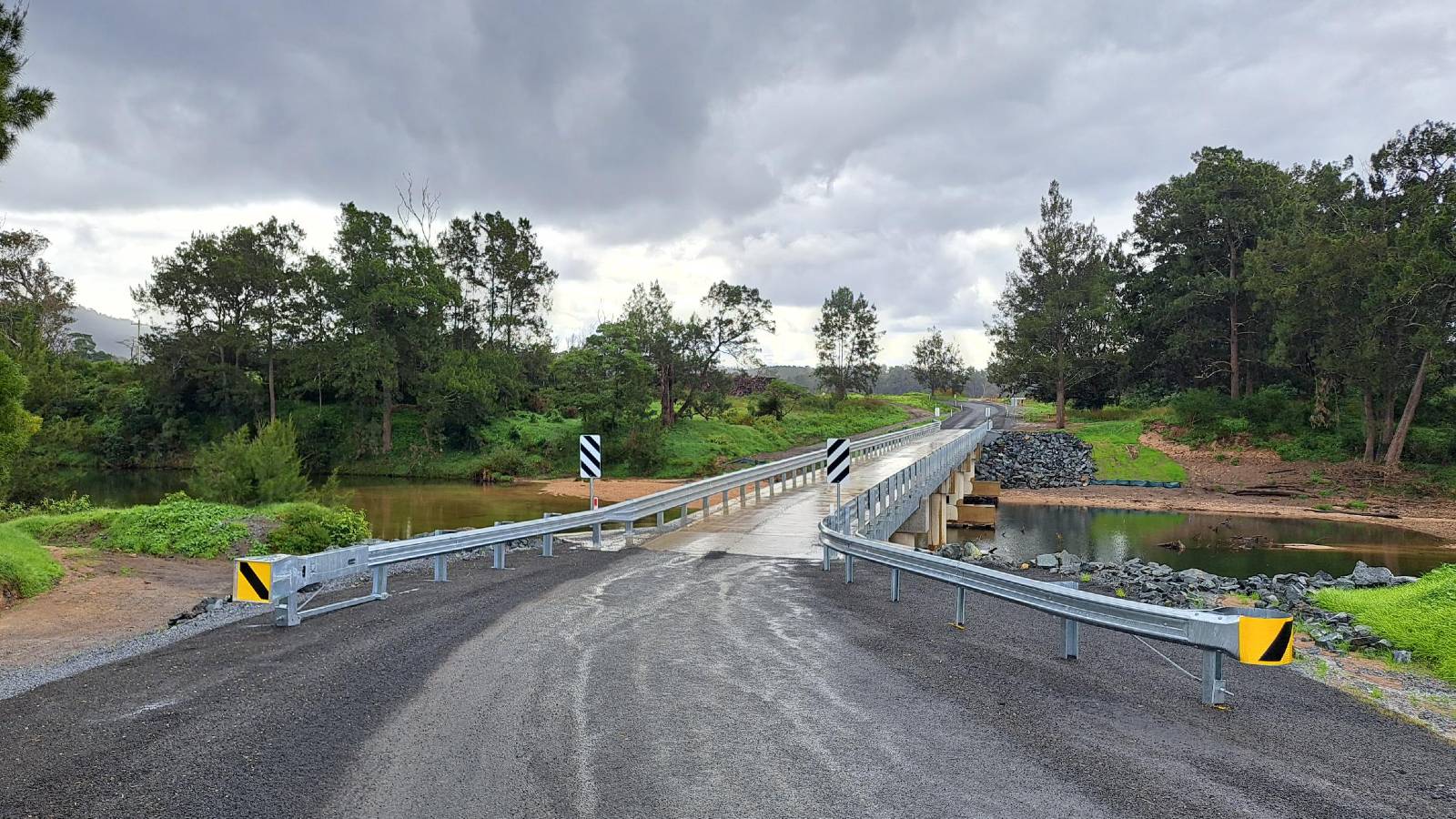 A sealed road leads to a long bridge lined with guardrail in a rural setting.
