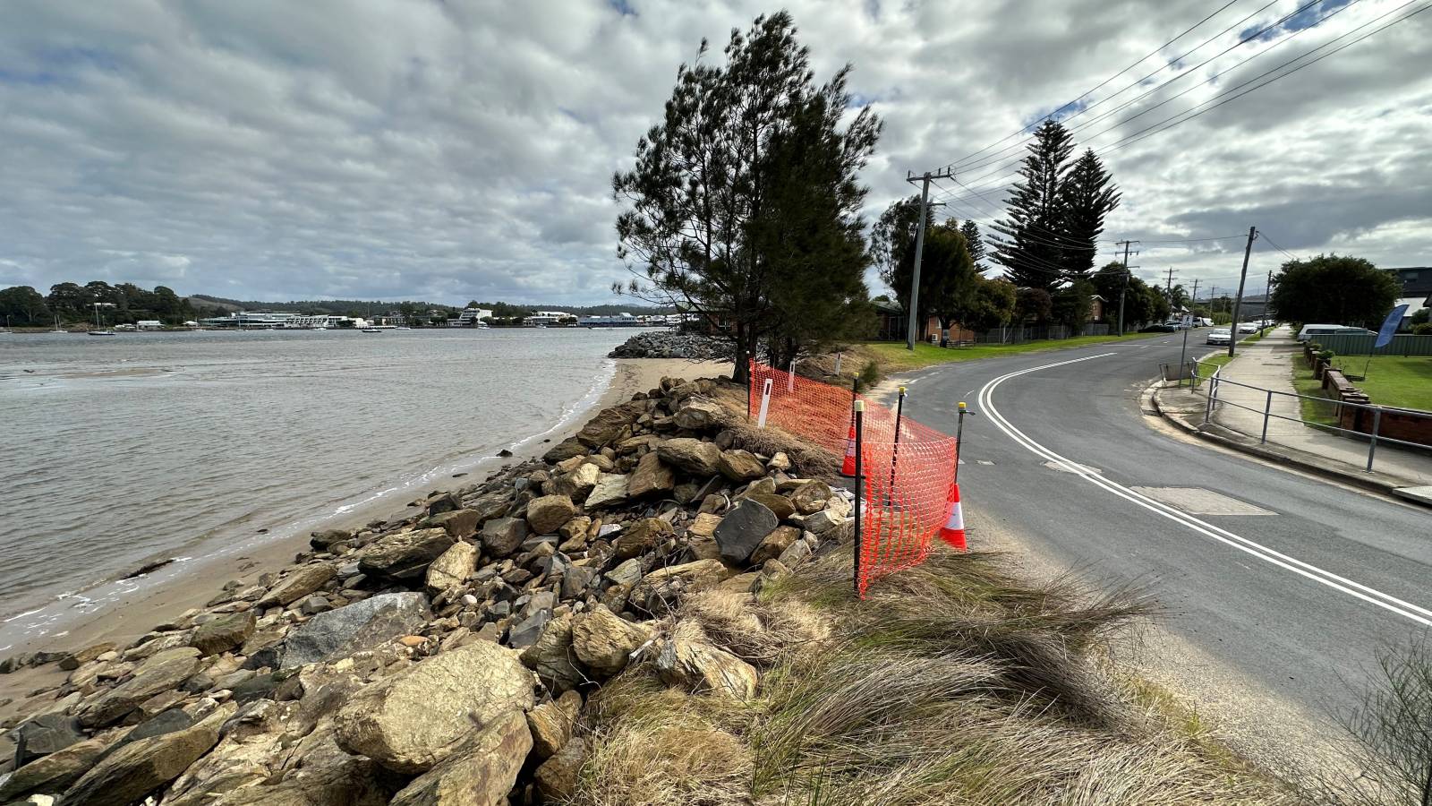 A rock seawall between the beach and road with barrier mesh along the roadside.