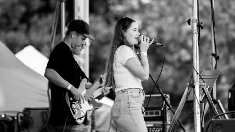 Black and white image of a girl singing on stage into a microphone with a guy behind her playing guitar.