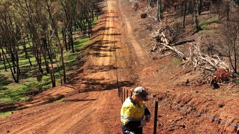 A woman rebuilds a rural fence in a burnt hilly landscape.