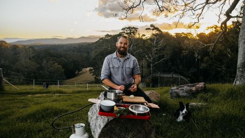 Bearded man sitting in paddock with dog while cooking truffles over makeshift frypan