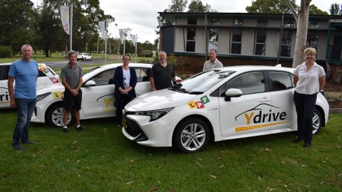 Six people stand in front of three cars with the Y drive logo