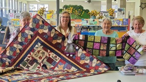 four older ladies standing behind a table and holding up a patchwork quilt