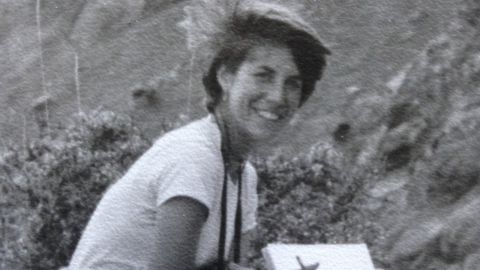 A black and white grainy image of a woman on a rocky hill with a pen and notebook.