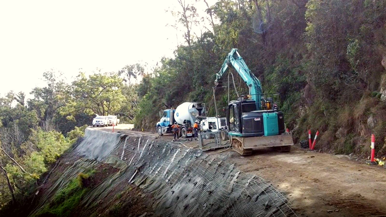 An excavator and a concrete agitator truck take up the whole of a dirt road while repairing the downhill slope.