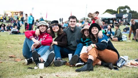 A group of young people sit on the grass at what looks like a festival.