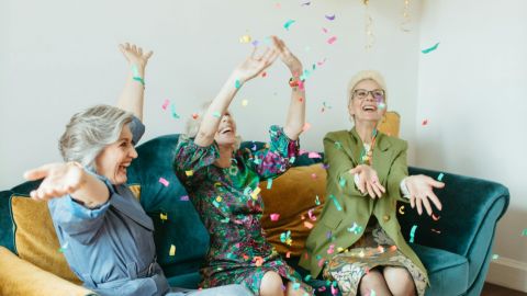 Three women sitting on a couch laughing through falling confetti.