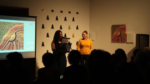 Two women stand in front of a crowd in a dimly lit room, next to an artwork on the wall and on a projector.