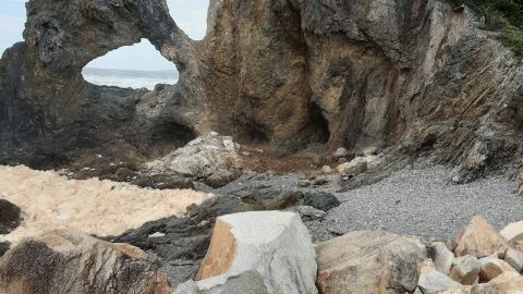 A rock formation with a hole shaped like Australia, next to a cliff, with piles of rocks  around it.